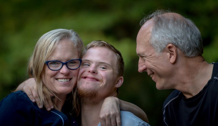 Glücklich strahlende Eltern und jugendlicher Sohn mit Downsyndrom umarmen sich vor grünem Hintergrund.
